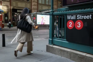 Person in beige coat with a black bag walking past a Wall Street subway sign and red circular number markers on a green railing.